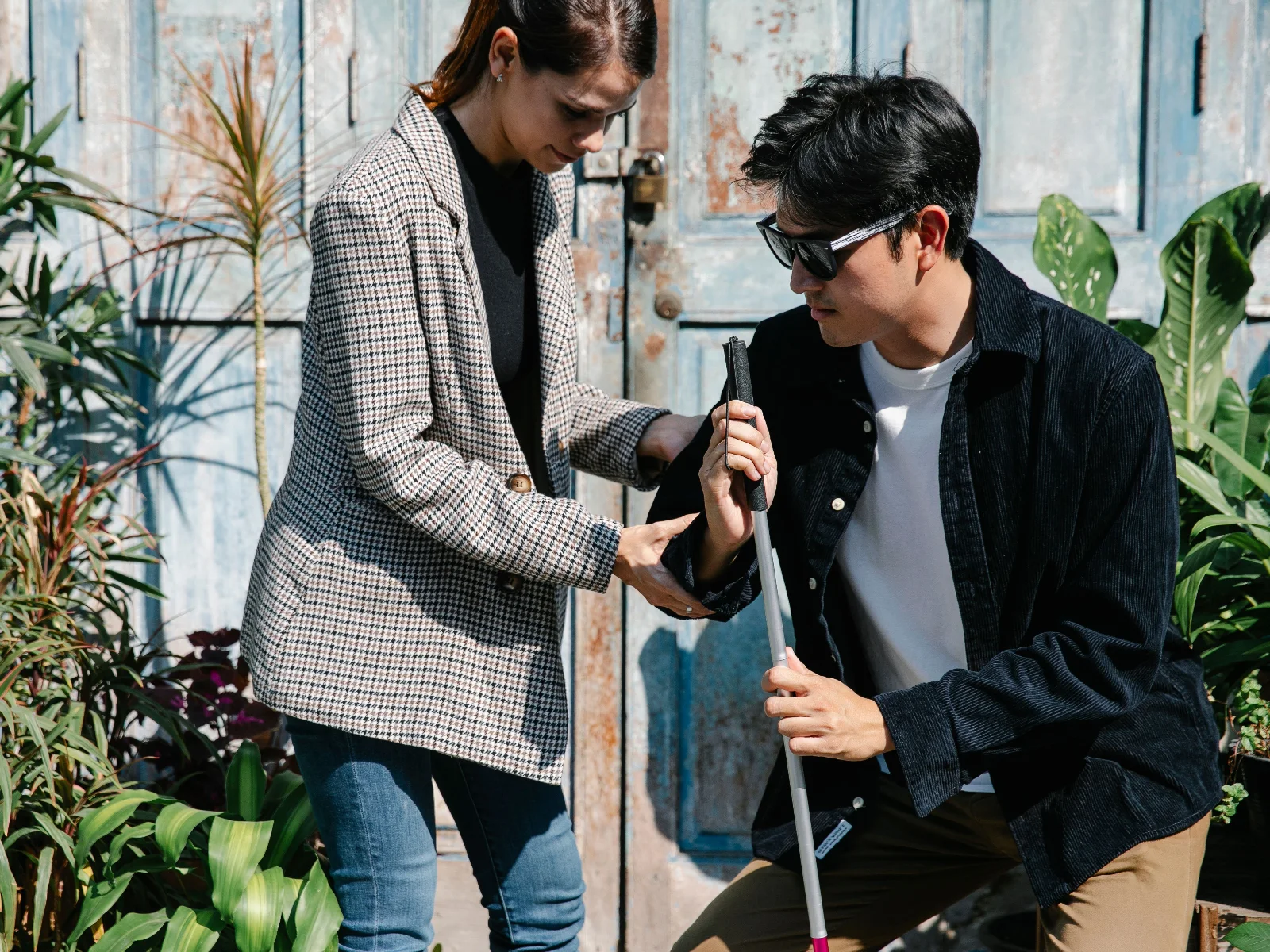 A woman walking alongside a man who uses a white cane, helping him find his way outdoors