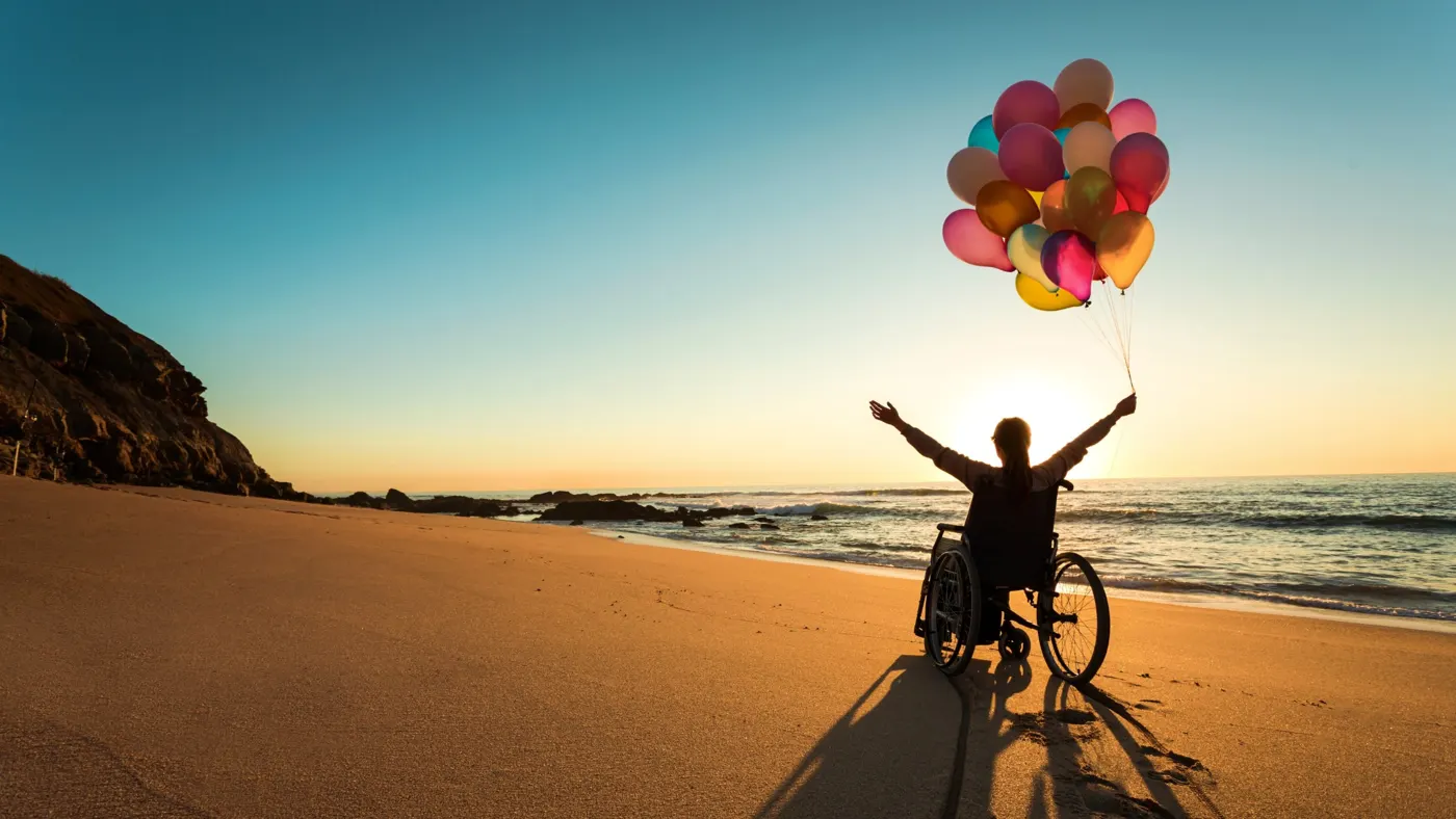 A woman in a wheelchair raising balloons on a beach at sunset
